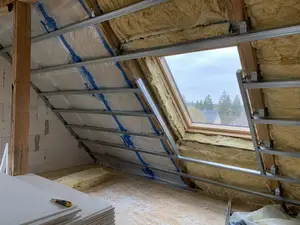 Poddasze w trakcie adaptacji z wełną mineralną między krokwiami, folią paroizolacyjną, metalowymi profilami stelaża g-k i oknem dachowym